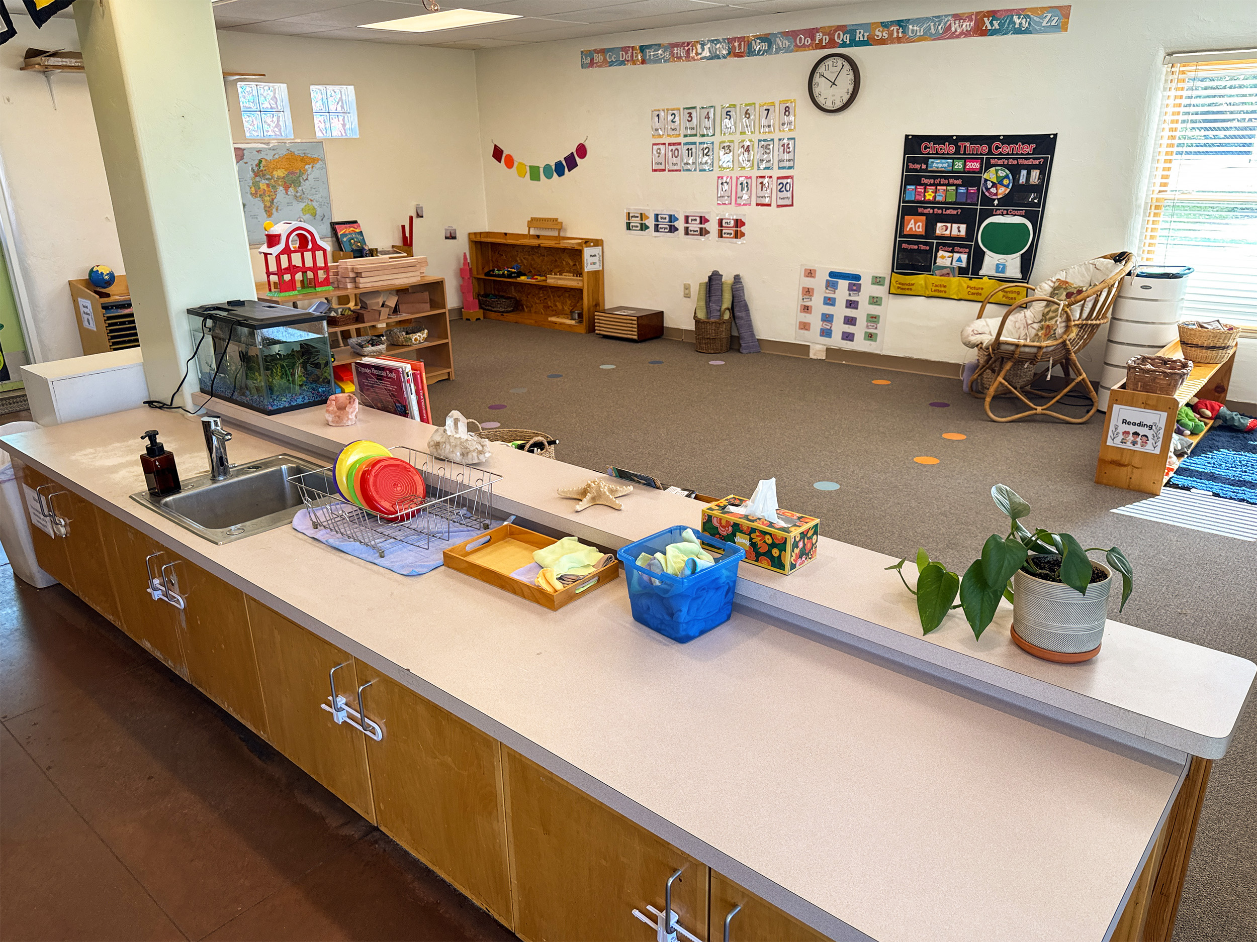 Image shows a wide shot of a classroom with a child-sized countertop and sink in the foreground. There are shelves and classroom materials in the background. 