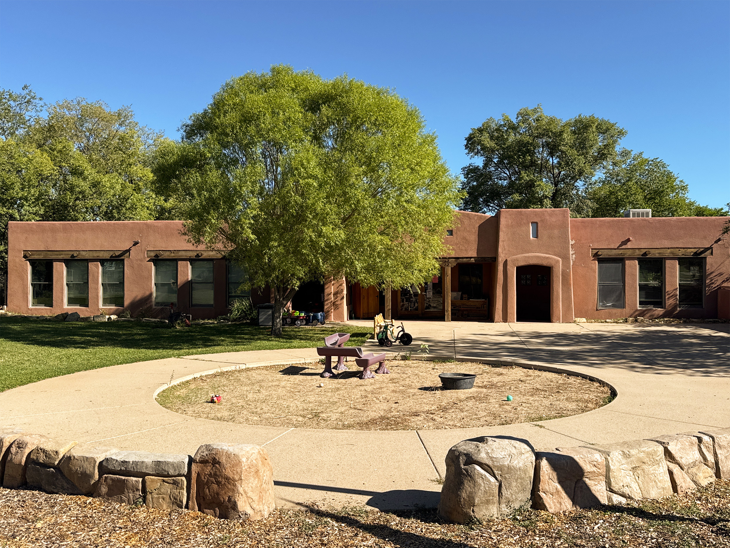 Image shows the front of the school with a large tree and a sand box with toys. 